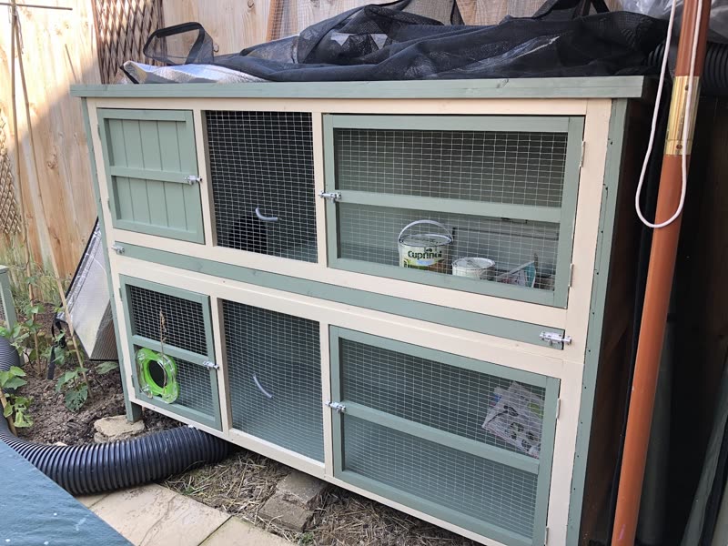 A two-storey wooden hutch with mesh doors and a weatherproof cover on the paved garden path at Dereham Little Pet Hotel